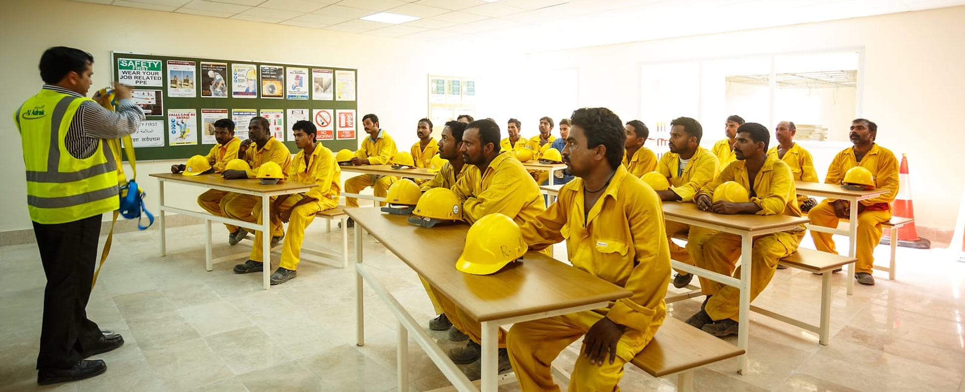 A safety induction at Al Adrak: an instructor presenting to a full room of the workforce in protective clothing, with the site safety noticeboard behind him